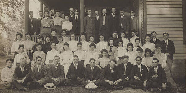 A large group of men and women in early 20th-century attire posed on a wooden porch.