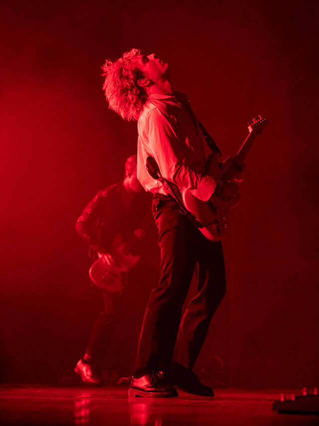 Two musicians performing on stage under red lighting, with one prominently playing a guitar.