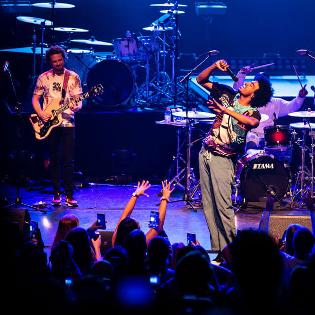 A singer and guitarist perform on stage with a drummer in the background, surrounded by blue and purple stage lighting, while the audience captures the moment on their phones.