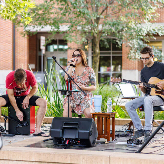 Outdoor music performance with a singer, a guitarist, and a percussionist.