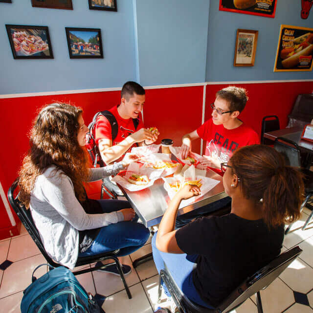Four people sitting at a table in a bright restaurant eating hot dogs.