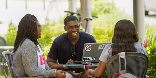 Three people sitting at a table outdoors, engaging in conversation, with a laptop in front of them.