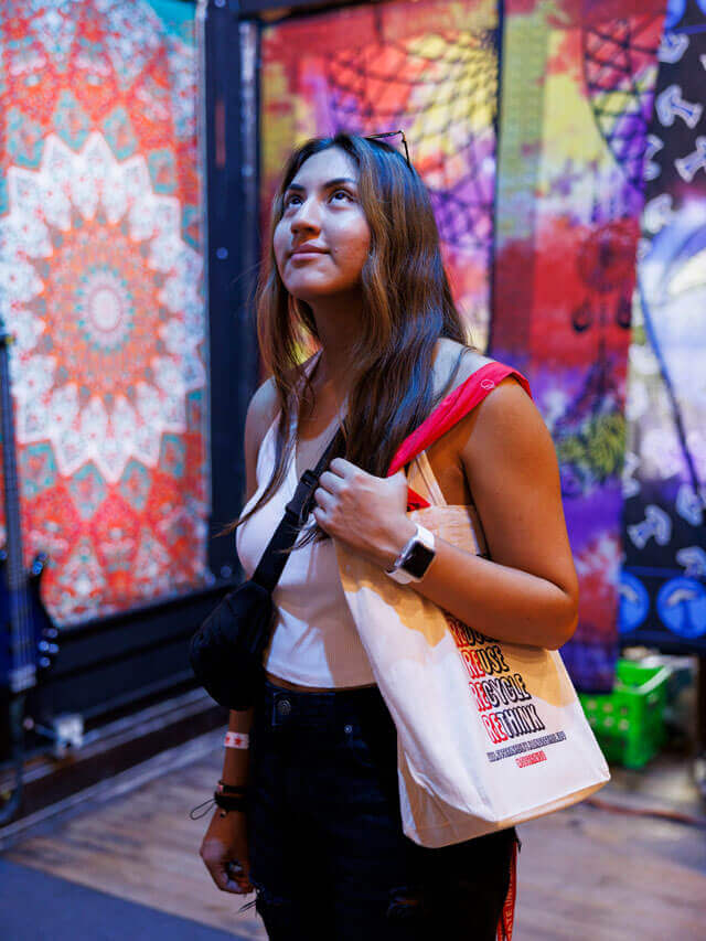 A person stands indoors, gazing upward, with colorful patterned tapestries in the background.