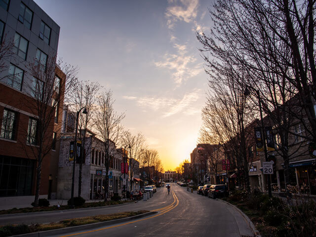 Urban street at sunset with bare trees, buildings, and a cyclist.