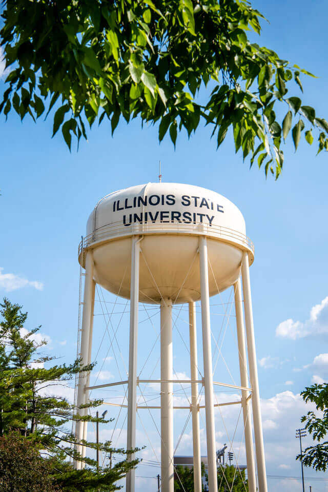 A white water tower labeled 'ILLINOIS STATE UNIVERSITY' against a blue sky, framed by green leaves.