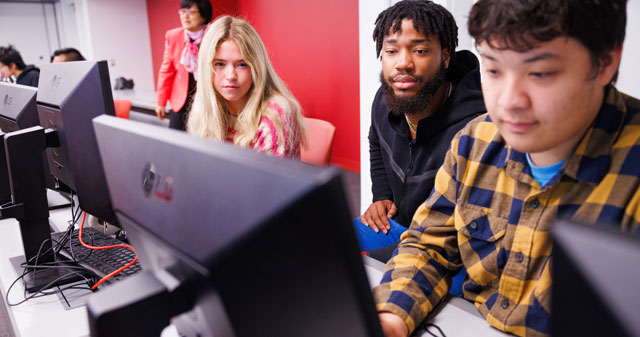 Three people working at computer stations in a classroom setting.