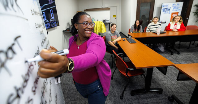 A woman in a pink sweater writes on a whiteboard as others watch in an office setting.