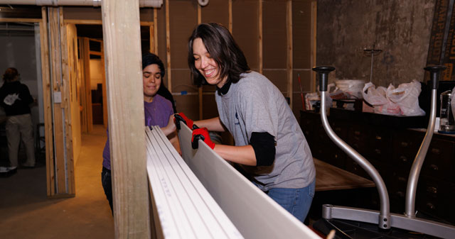 Two people carrying a piece of drywall in a construction room.